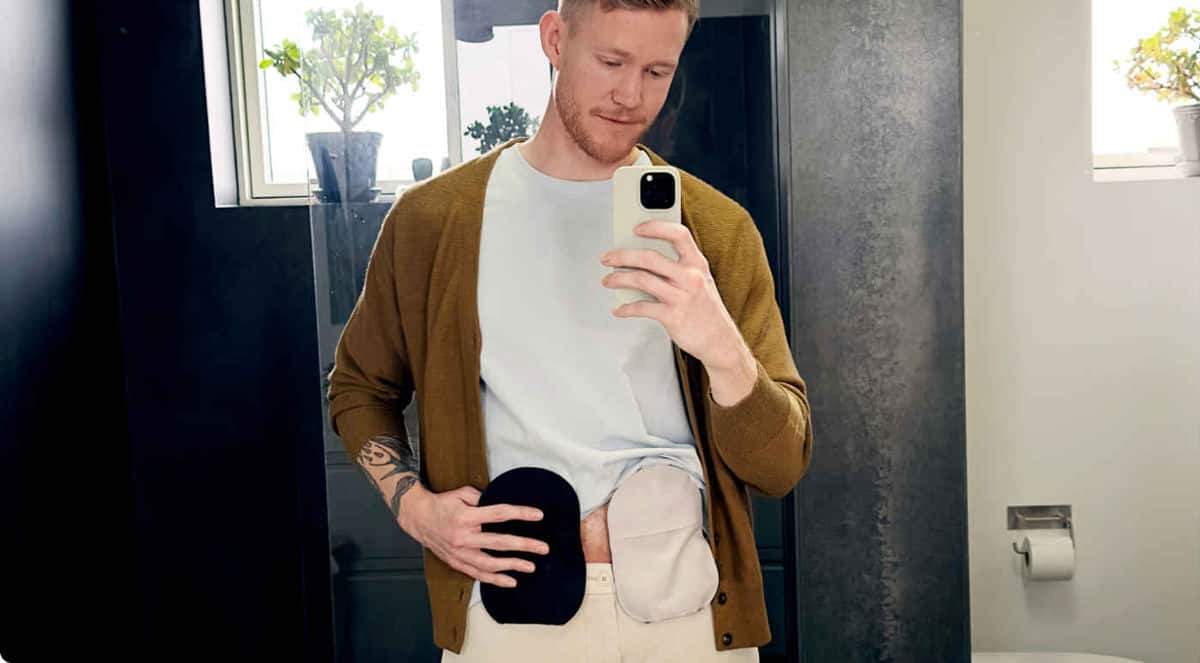 Man taking a picture in front of the mirror wearing a grey ostomy bag and a black ostomy bag