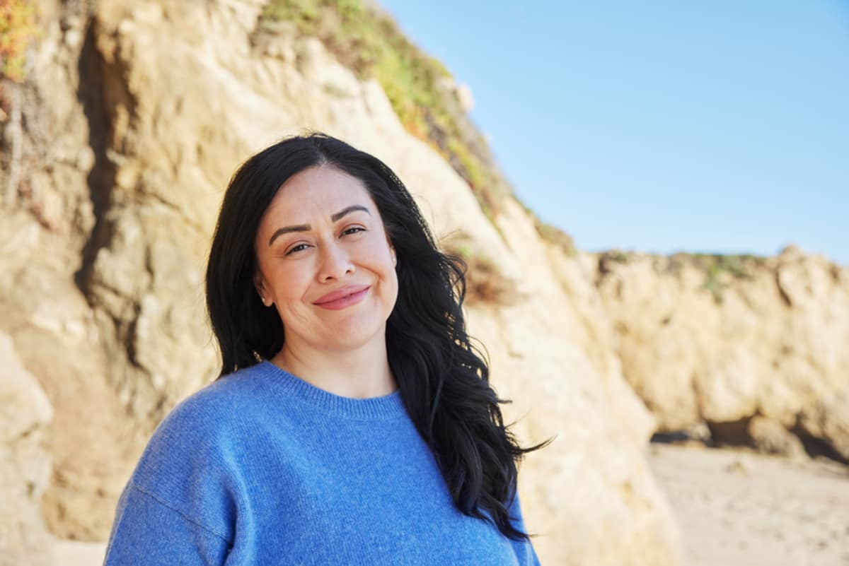 woman is stood at the beach and wearing a blue jumper, whilst smiling at the camera.